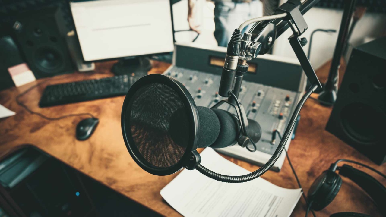 Close-up of a microphone in a professional voiceover studio, surrounded by a mixing console, monitor, keyboard, and papers scattered on the wooden desk in the background.